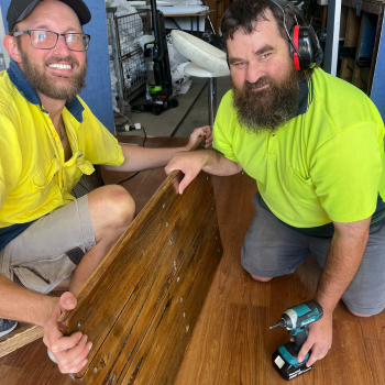 Two workers kneeling together, smiling, beside a restored wooden bench and a power drill.